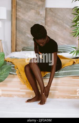 Bella donna afroamericana con dreadlocks seduta sul letto in studio decorato e massaggiante piede a mano. Persona nera che ha lesioni sportive, muscolo Foto Stock