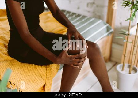 Afro american donna seduta e massaggiante ginocchiera da mani primo piano, fuoco selettivo. Persona nera che ha lesioni sportive, sforzo del muscolo della gamba. Peloso e. Foto Stock