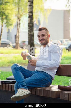 Un uomo serio siede su una panca, beve caffè, tiene dollari. Giovane uomo sullo sfondo di auto passanti, caldo giorno d'estate soleggiato. Luce calda e soffusa, primo piano. Foto Stock