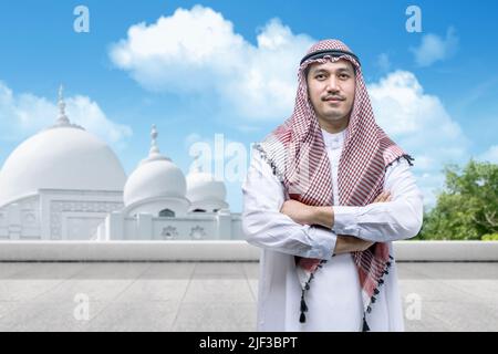 Uomo arabo che indossa un keffiyeh in piedi con braccia ripiegate con moschea e sfondo blu cielo Foto Stock