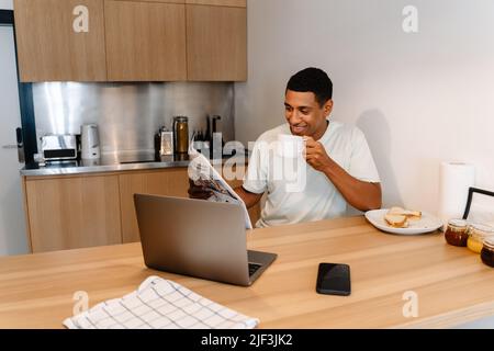 Uomo nero che legge il giornale e beve il caffè mentre lavora con il computer portatile in hotel Foto Stock