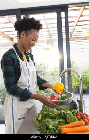 Sorridente donna afroamericana di metà adulta che indossa grembiule pulizia verdure in cucina lavandino Foto Stock