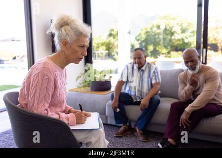 Terapista femminile multirazziale con appunti guida anziani in sessione di terapia di gruppo in casa di cura Foto Stock