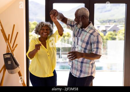 Allegro uomo anziano afroamericano che fa la salsa ballare con la donna contro la finestra in casa di cura Foto Stock