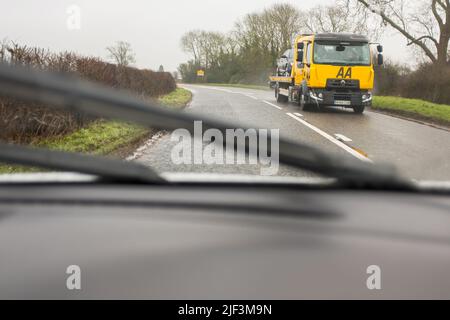 Auto caricata su un camion di recupero di guasti AA su una strada di campagna, vista del parabrezza da un'auto in arrivo, in una giornata piovosa. Foto Stock