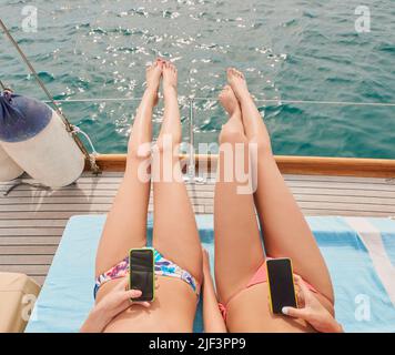 Due amici che prendono il sole sulla barca durante la crociera di festa insieme usando i telefoni cellulari per inviare messaggi di testo. Due donne che si trovano sulla barca utilizzando smartphone e. Foto Stock
