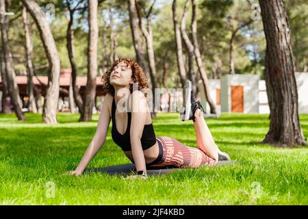Bella donna rossa che indossa abiti sportivi in piedi sul parco cittadino, all'aperto che fa yoga, stretching su tappeto yoga. Esercitarsi a stretching alto cobra A. Foto Stock