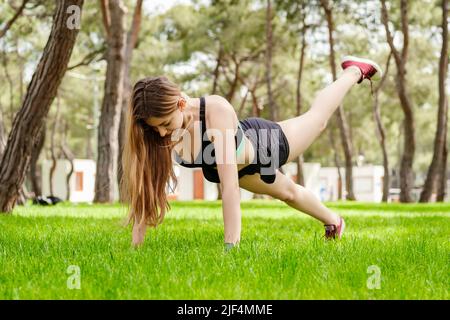 Giovane bella donna che indossa abiti sportivi sul parco cittadino, donna all'aperto che fa push up con una gamba su. Sport all'aperto, concetti di vita sana. Foto Stock