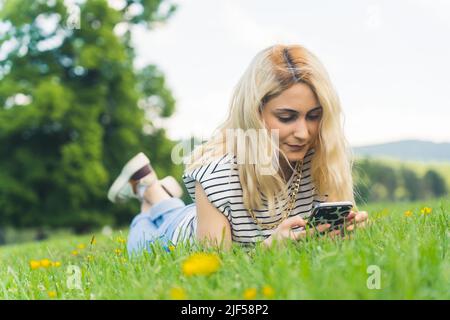 Ragazza caucasica bionda che riposa sull'erba e controlla le reti sociali. Pieno colpo all'aperto. Foto di alta qualità Foto Stock