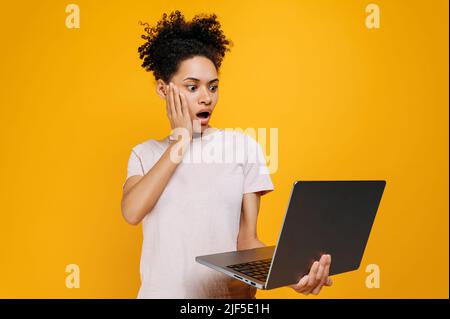 Scioccata stupita giovane afroamericana con capelli ricci, tiene un portatile aperto in mano, guarda lo schermo in sorpresa, ha visto informazioni inaspettate, si erge su isolato sfondo arancione Foto Stock