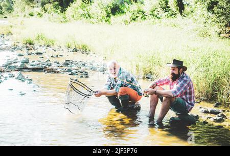 Squadra di pesca. Uomini brutali seduti vicino al fiume acqua. Continuare a pescare. Divertimento e relax. Pescatore che cattura il pesce. Buon tempo. Rilassatevi nella natura Foto Stock