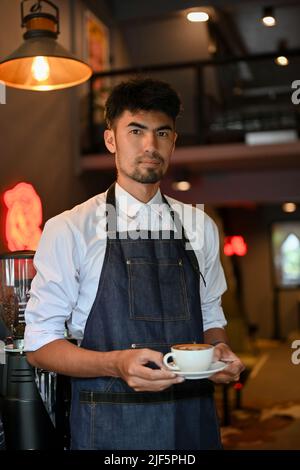 Bella e professionale barista maschile asiatico o cameriera con grembiule che serve una tazza di caffè, in piedi nella moderna caffetteria. Foto Stock