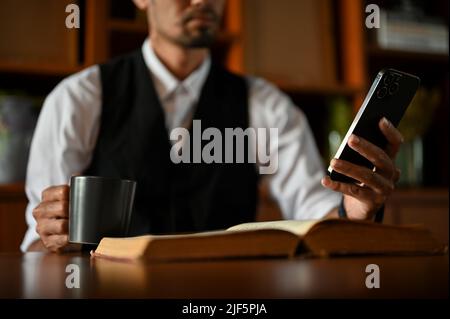 Un uomo d'affari o un CEO maschio di successo alla sua scrivania utilizzando uno smartphone per controllare la sua e-mail e sorseggiare un caffè caldo al mattino. Immagine ritagliata, primo piano Foto Stock