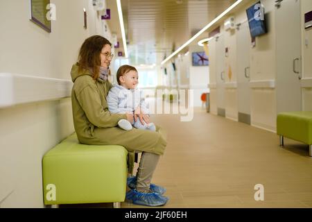 Donna madre con bambino ragazzo nelle sue braccia visita nella clinica sala. Mamma con un bambino nel corridoio dell'ospedale in attesa di un medico nominare Foto Stock