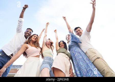 Vista ad angolo basso di un gruppo di amici multietnici che alzano le mani e ridono mentre si posano insieme all'aperto. Concetto di amicizia. Foto Stock