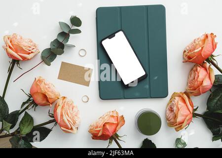 Smartphone a schermo vuoto con tablet, fiori, verde e biglietto da visita su bianco, vista dall'alto, disposizione piatta. Modello di lavoro femminile creativo, social network Foto Stock