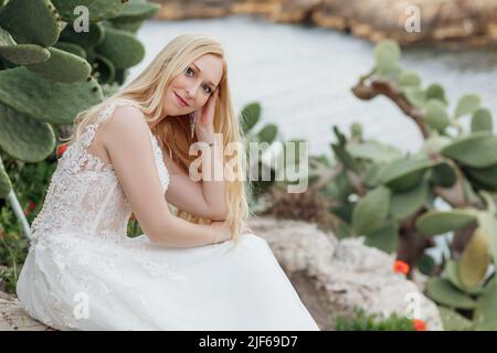 Ritratto di giovane sorridente donna attraente sposa seduta su roccia vicino verde enorme foglie di opuntia cactus. Matrimonio. Foto Stock