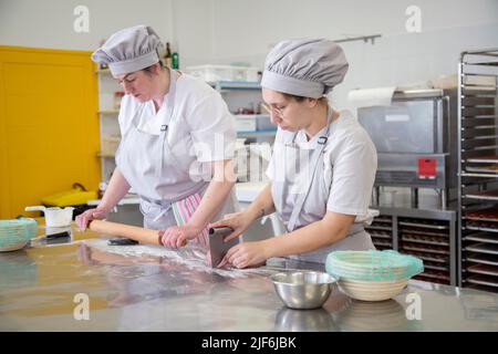 Donne focalizzate in uniformi arrotolando e tagliando pasta cruda su tavola metallica mentre si preparano per la cottura nella scuola di panetteria leggera Foto Stock