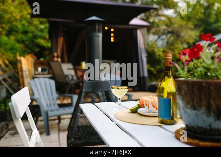 Bicchieri con champagne serviti su un tavolo di legno bianco con gamberi e bottiglia sulla terrazza dell'edificio con alberi su sfondo sfocato Foto Stock