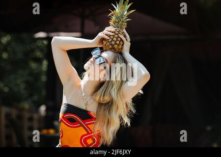 Giovane donna in elegante top e occhiali da sole con ananas maturo sulla testa, mentre in piedi sulla terrazza dell'edificio Foto Stock