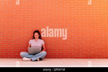 Corpo completo di studentessa femminile concentrata in abbigliamento casual surf internet su netbook mentre si siede sul pavimento vicino muro di mattoni Foto Stock