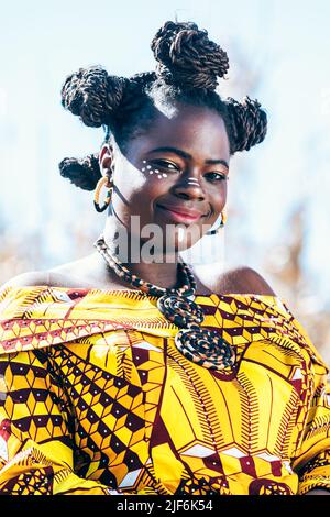 Giovane donna nera in vivace autentico vestito africano con ciambelle e trucco tradizionale ripiegando le mani sul ventre e guardando la macchina fotografica mentre in piedi Foto Stock