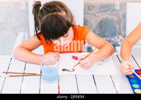 Ragazza focalizzata pittura su carta con pennello al tavolo con tavolozza di acquerelli e vetro d'acqua mentre si siede sulla terrazza dell'edificio Foto Stock