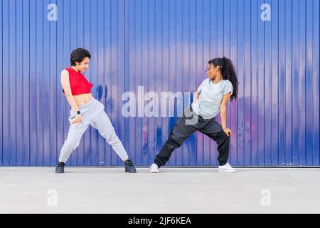 Vista laterale completa del corpo di giovani amici multirazziali che ballano sulla strada vicino al muro blu della città Foto Stock