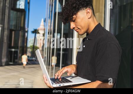 Vista laterale di etnia maschile freelancer in polo nera appoggiata sul muro e lavorando a distanza su un moderno computer portatile mentre si trova in città strada Foto Stock