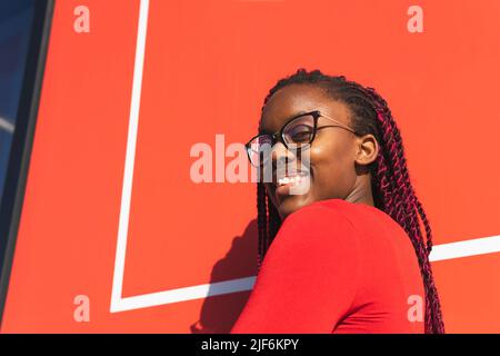 Vista laterale della giovane donna afroamericana in abiti alla moda e occhiali che guardano la macchina fotografica mentre si trova su sfondo rosso Foto Stock