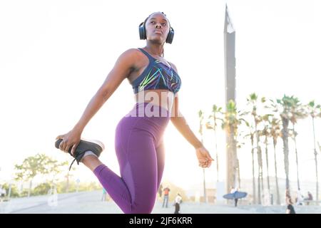 Vista laterale dell'atleta femminile in abbigliamento sportivo e cuffie che allungano le gambe durante il riscaldamento prima dell'allenamento Foto Stock