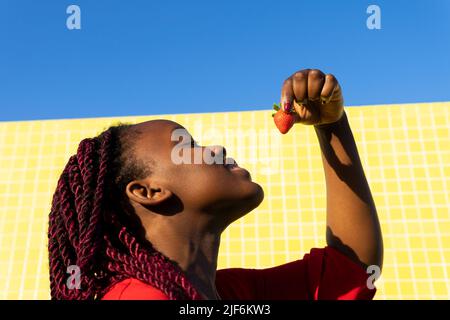 Da sotto allegra giovane afroamericana femmina in abito rosso mangiare fragole su sfondo giallo in una giornata di sole Foto Stock