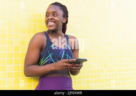 Indossa un abbigliamento sportivo femminile nero utilizzando il telefono in piedi all'aperto su sfondo giallo Foto Stock