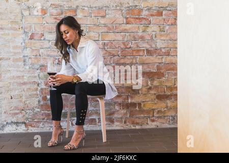 Donna adulta premurosa in abiti casual eleganti con bicchiere di vino rosso con lunghi capelli seduti sullo sgabello contro la parete di mattoni Foto Stock