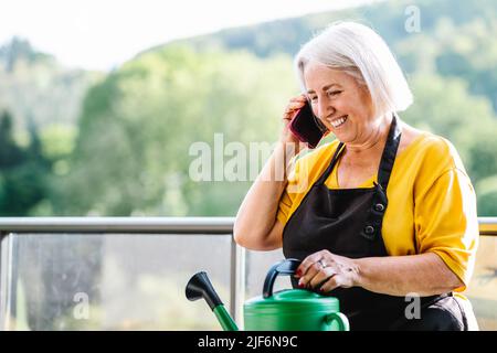 Donna anziana positiva in abiti casual riposati dopo annaffiare piante e parlare al telefono cellulare Foto Stock