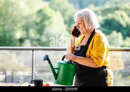 Donna anziana positiva in abiti casual riposati dopo annaffiare piante e parlare al telefono cellulare Foto Stock