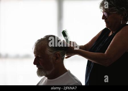 Vista laterale della moglie di raccolto messa a fuoco negli occhiali con pettine che fa l'acconciatura all'uomo anziano bearded nella stanza luminosa con le finestre Foto Stock