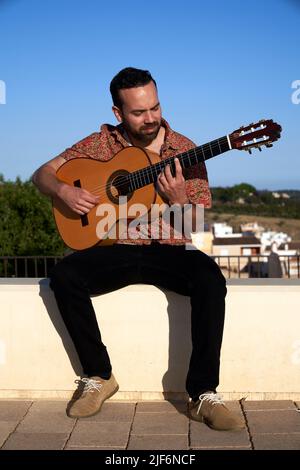 Corpo pieno di musicista bearded che suona la canzone sulla chitarra acustica mentre si siede con gli occhi chiusi sul bordo contro il cielo blu nuvoloso nella giornata di sole in città Foto Stock