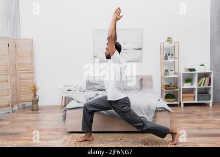 vista laterale dell'uomo afroamericano che pratica yoga in posa di guerriero e meditating in camera da letto Foto Stock