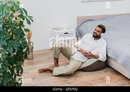happy african american uomo in cuffie wireless ascoltare musica mentre si tiene lo smartphone e sedersi vicino a letto Foto Stock