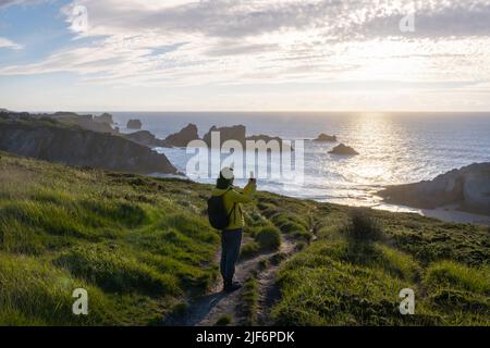 Vista posteriore di una donna escursionista che fotografa la scogliera utilizzando smartphone vicino al mare contro il cielo Foto Stock