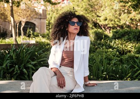 Donna d'affari ispanica in tuta bianca alla moda e occhiali da sole con capelli ricci seduta su panca di pietra e guardando lontano mentre spende la giornata estiva soleggiata dentro Foto Stock