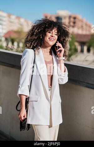 Buon imprenditore ispanico femmina in abito bianco con capelli ricci sorridenti e parlando su smartphone mentre si cammina vicino alla recinzione in giornata di sole sulla strada della città Foto Stock