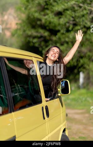 Una donna ispanica ottimista sorridente a occhi chiusi e che gode di vento mentre si sbirciava dalla finestra del RV durante un viaggio in natura il giorno d'estate Foto Stock