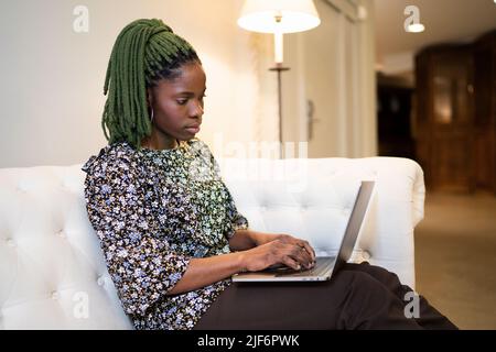 Giovane freelance afro-americana che digita su netbook moderno mentre lavora da remoto in salotto a casa Foto Stock
