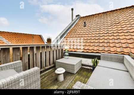 Comodo divano in vimini e poltrona in rattan posto sulla veranda di tipico edificio residenziale con tetto piastrellato e finestre su mansarda contro il blu nuvoloso Foto Stock