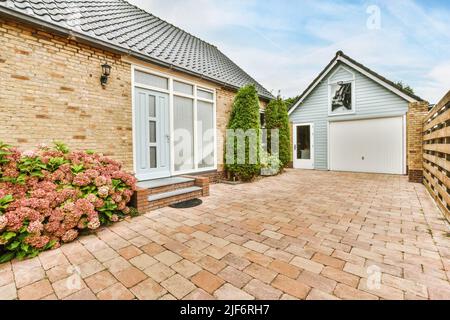 Casa residenziale con porta di vetro vicino patio con passerella lastricata e fiori in crescita e alberi in campagna il giorno d'estate Foto Stock