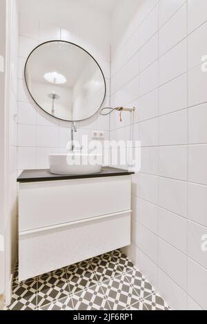 Lavarsi sopra i cassetti a parete con specchio posizionato su piastrelle bianche in un bagno elegante e leggero a casa Foto Stock