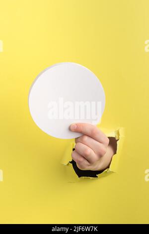 il bambino passa attraverso il foro in carta gialla con i bordi strappati che reggono un blocco di legno rotondo bianco, spazio di copia Foto Stock