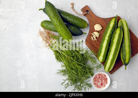 Cetrioli freschi sbucciati e tagliati su un tagliere di legno marrone con un mazzetto di aneto, aglio e sale rosa Foto Stock